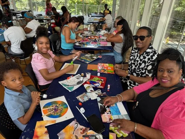 A group of children and adults sitting around tables in a brightly lit room engaged in arts and crafts activities, with various craft supplies on the tables.