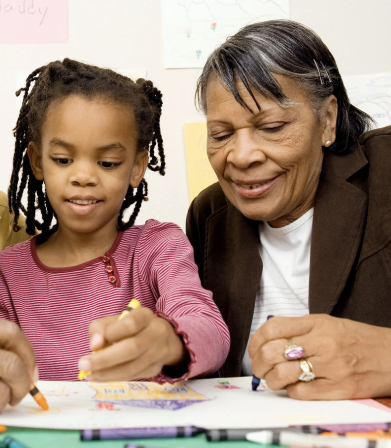 An elderly woman with dark hair and a brown blazer helping a young girl with braided hair during arts and crafts activity at a table, both smiling.