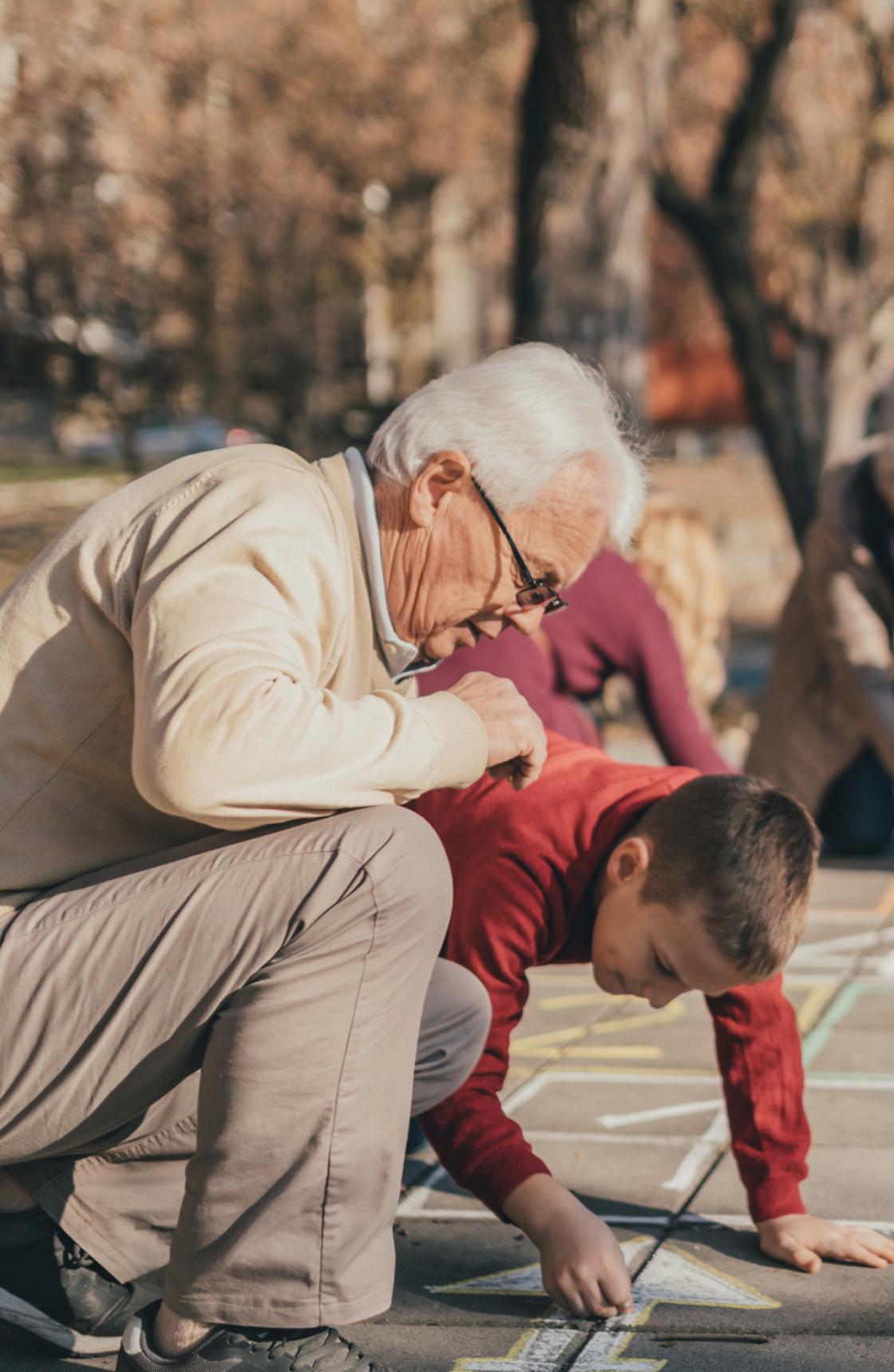An elderly man with glasses and a young boy drawing on the pavement with chalk during daytime, outdoors with trees and other people in the background.