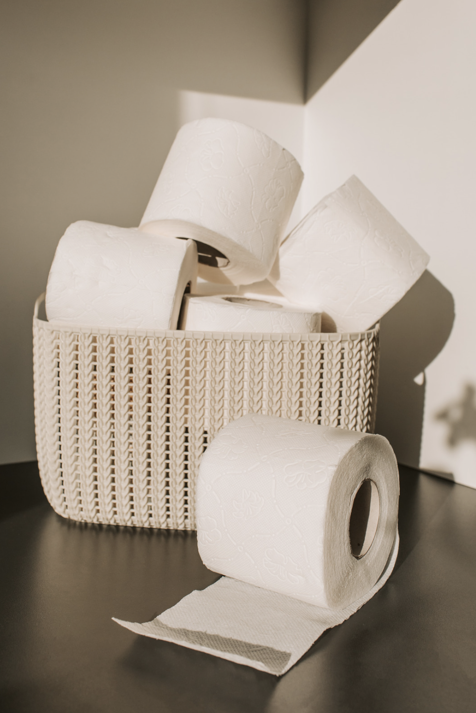 A white wicker basket filled with rolled toilet paper rolls, with one roll unspooled in front on a dark surface.