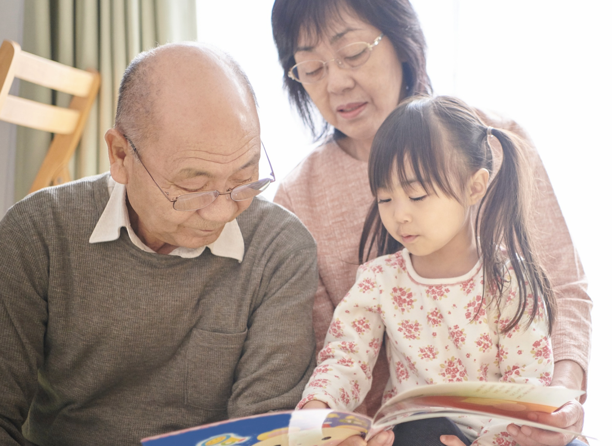 An elderly man and an elderly woman sitting with a young girl, reading a colorful book together indoors near a window with natural light.