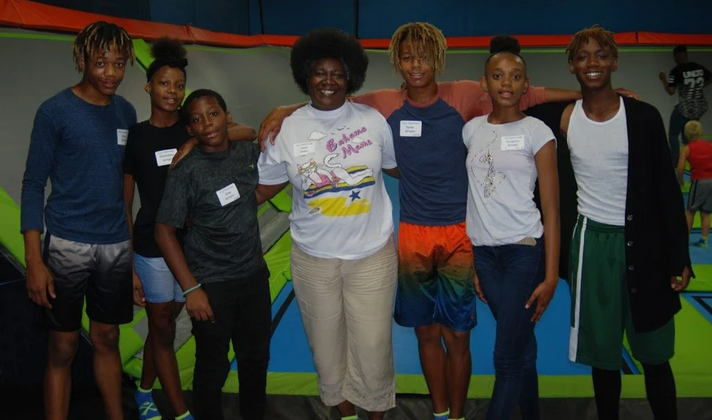 Group of seven children and a woman standing together inside an indoor trampoline park, smiling at the camera.