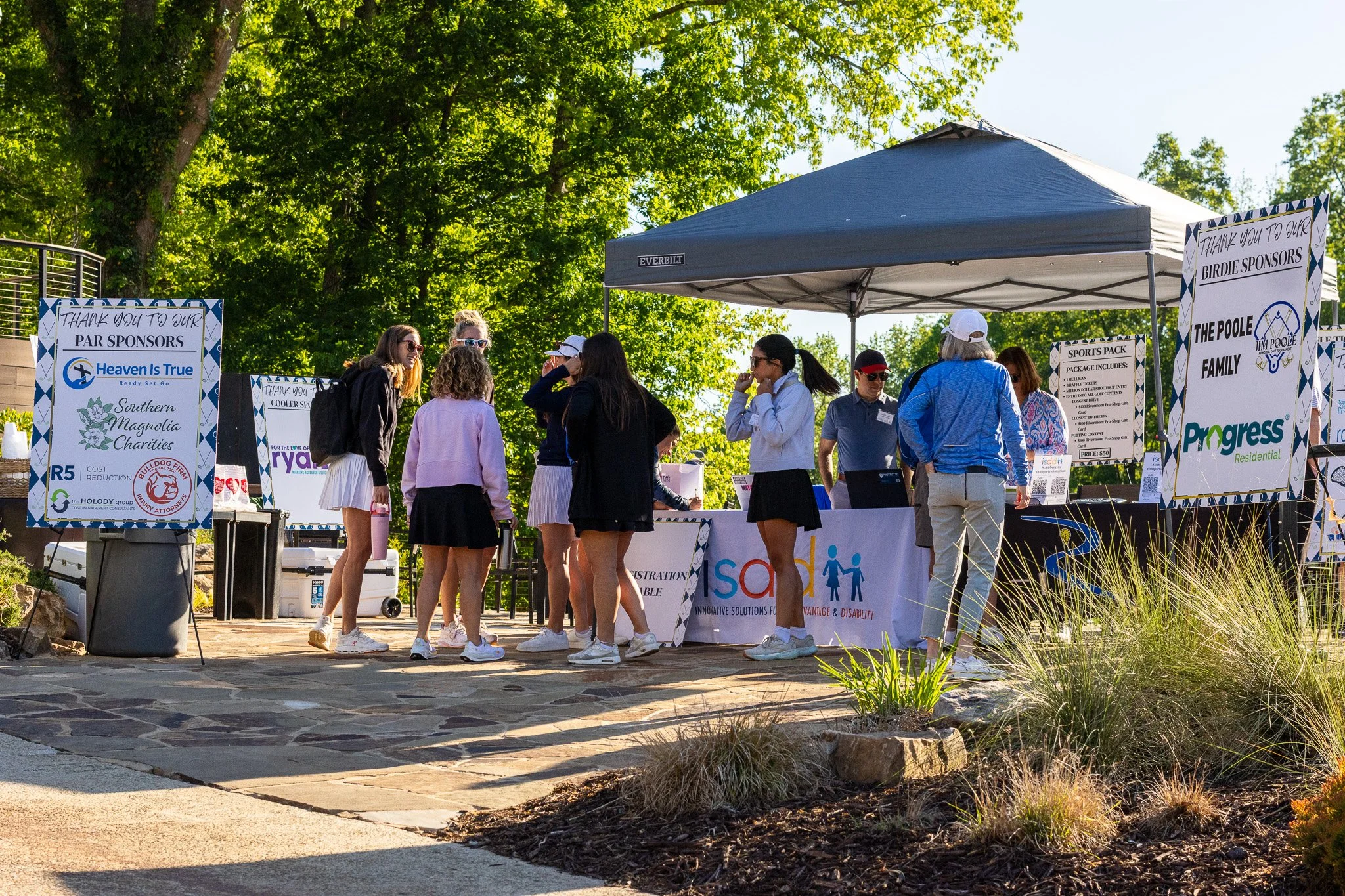 Group of people gathering around a booth with signs and banners at an outdoor event, with trees in the background and a large blue canopy tent overhead.