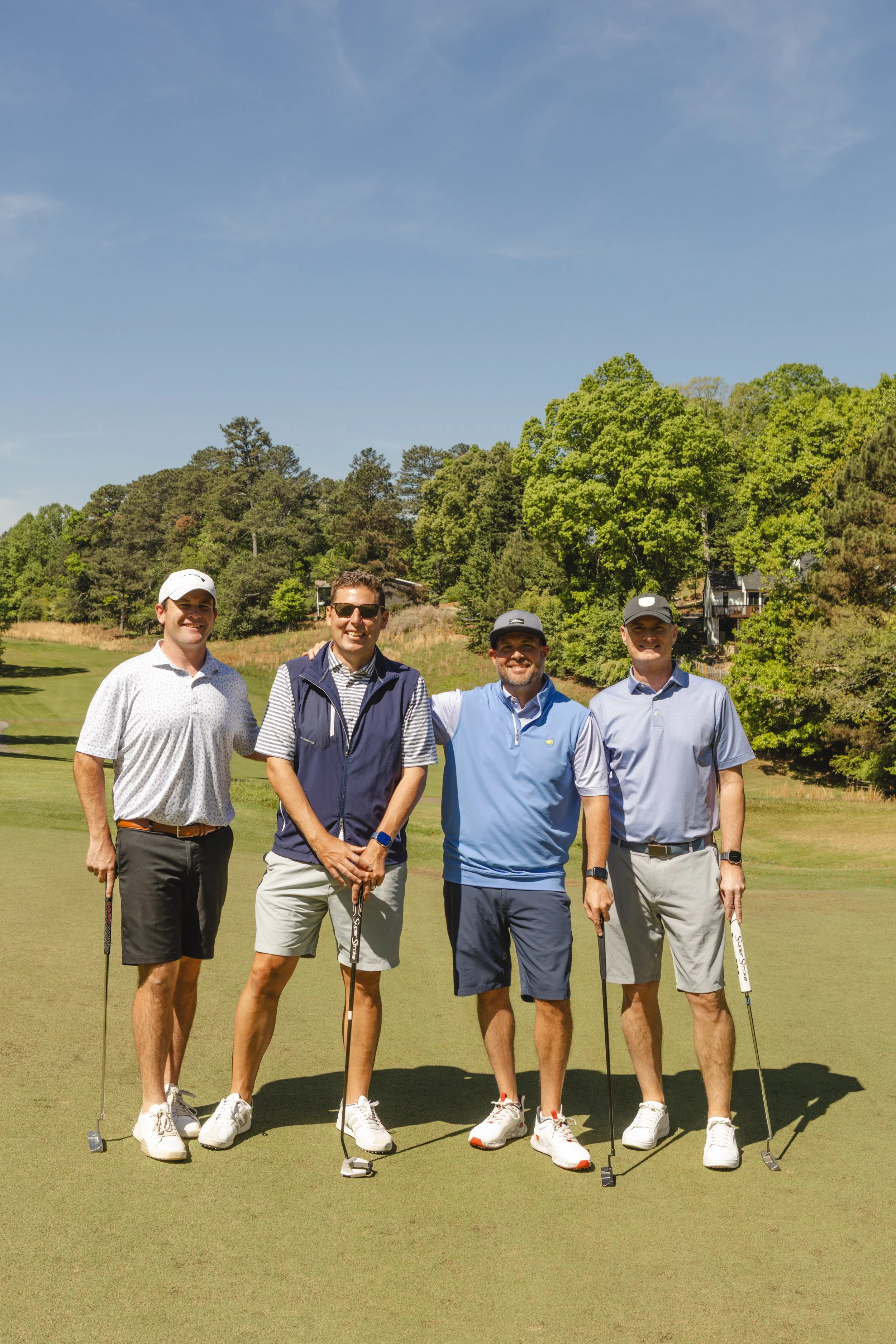 Four men standing on a golf course green, holding golf clubs, and smiling at the camera, with trees and a house in the background under a blue sky.