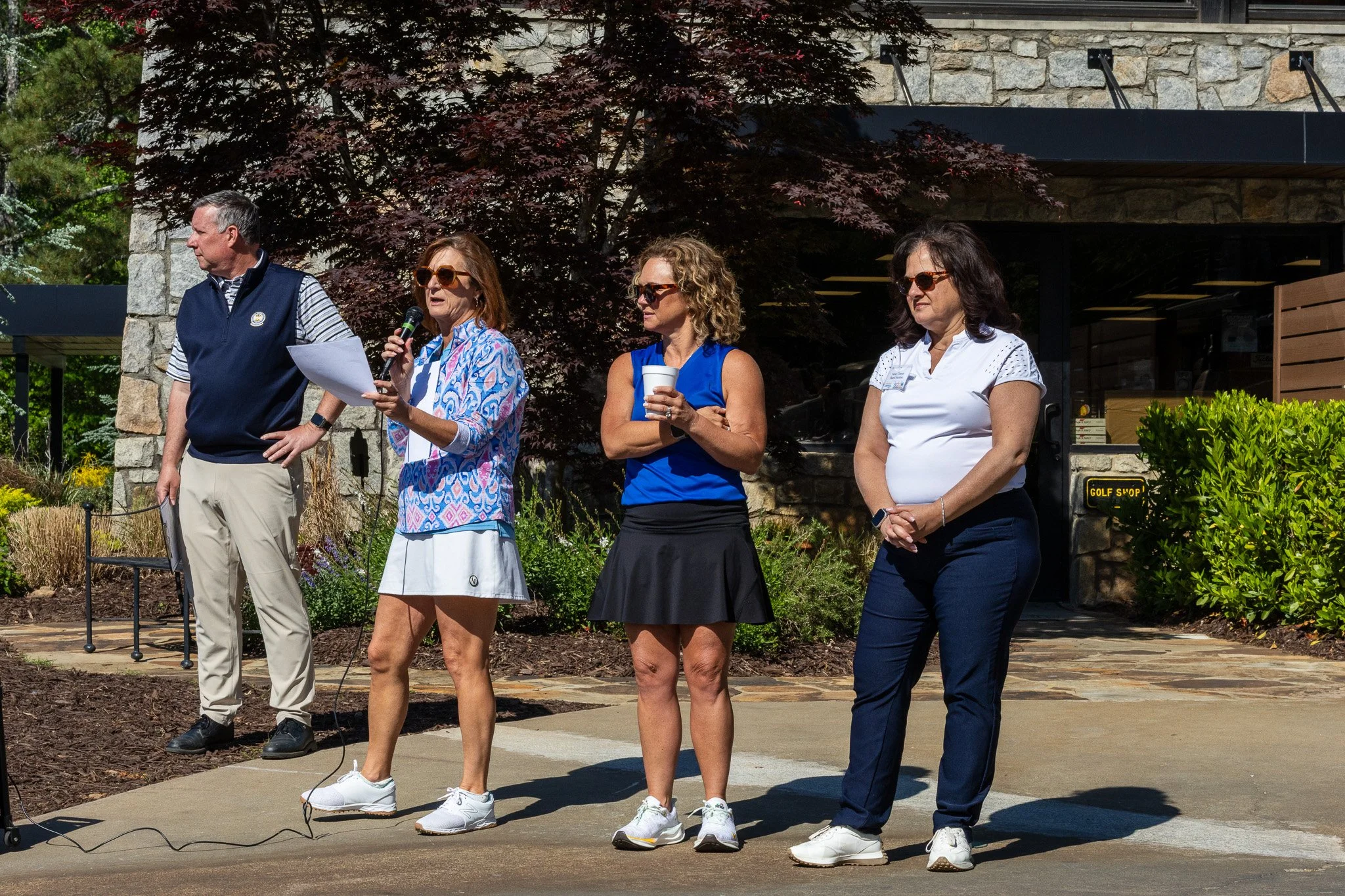 Four women and one man standing outdoors in front of a stone building and trees, some wearing sunglasses, with one woman speaking into a microphone and holding papers.