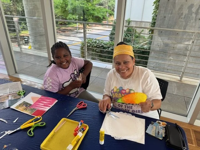 Two girls sit at a table with craft supplies, smiling, with large scissors, glue sticks, and paper in front of them.
