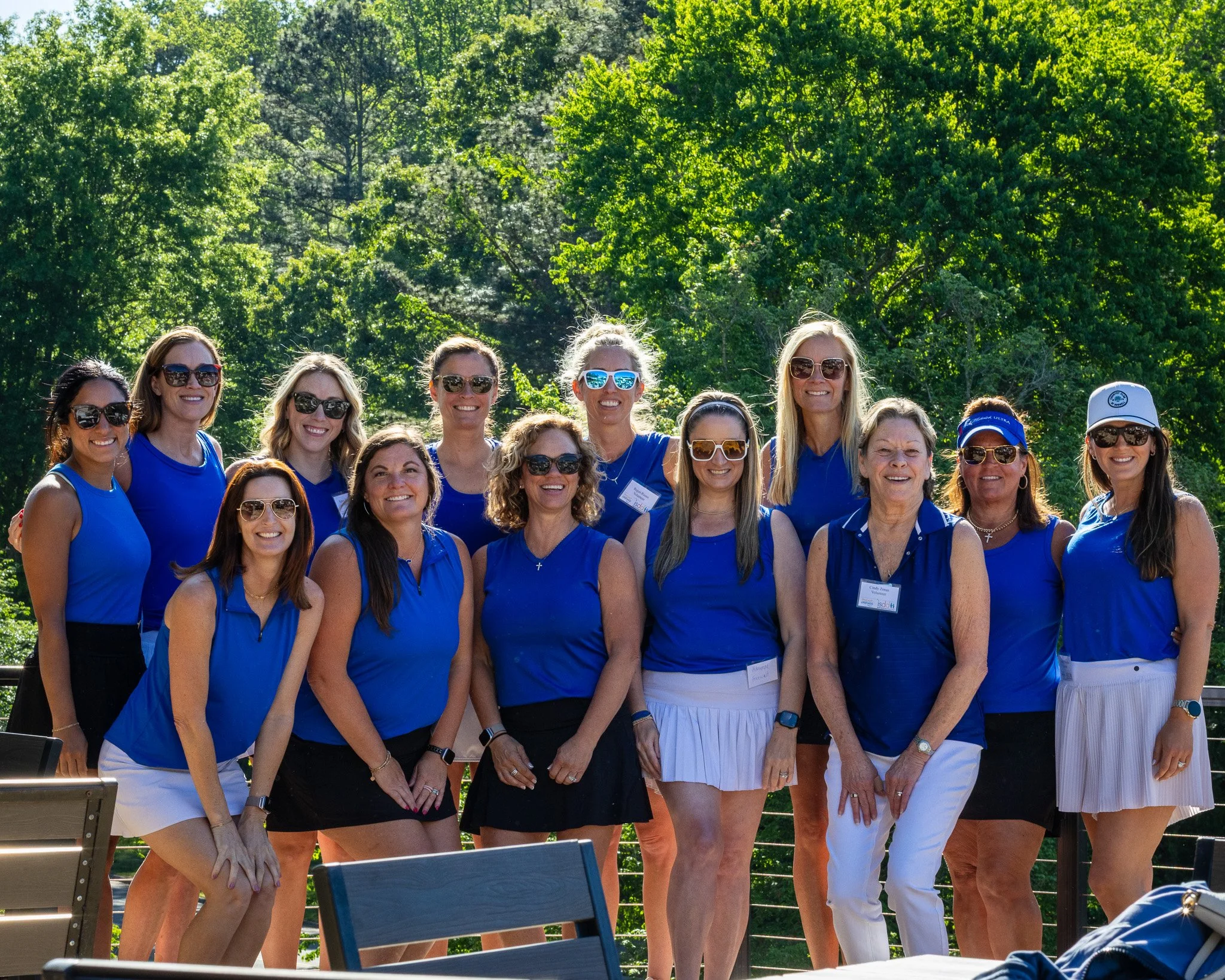 Group of women wearing blue shirts and sunglasses, standing outdoors with trees in the background.