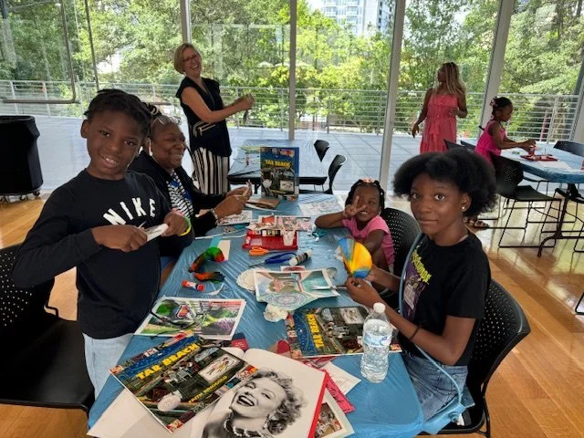 Children and adults gathered around a table with balloons, craft supplies, and posters, in a bright indoor space with large windows and trees outside.