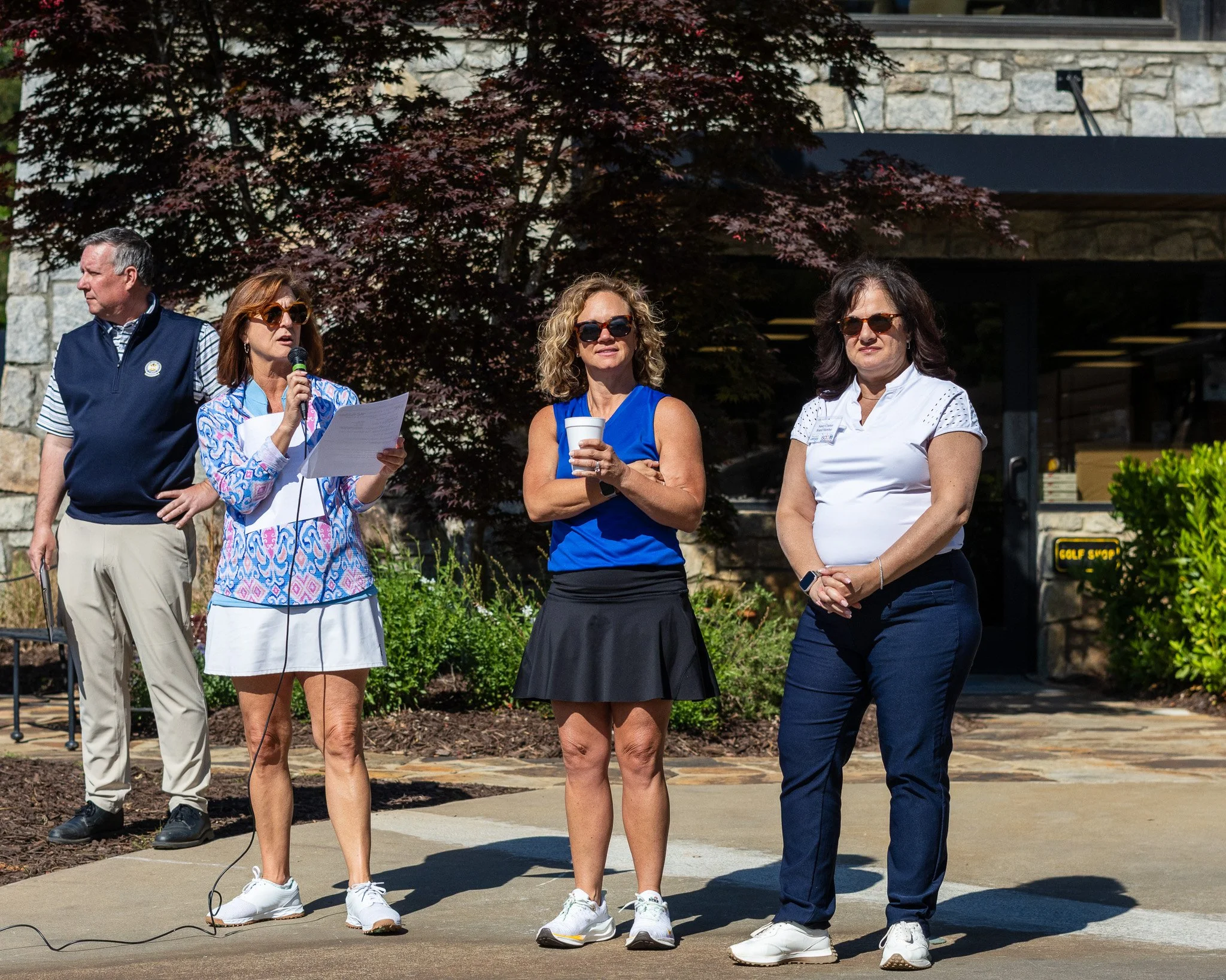 Group of five adults standing outdoors, participating in a speaking or presentation event, with a woman holding a microphone and paper, in front of a tree with reddish leaves.
