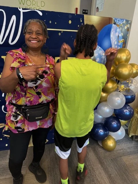 A woman smiling and pointing at a child from behind decorated with a balloon display of gold, silver, and blue balloons, in front of a dark blue backdrop.