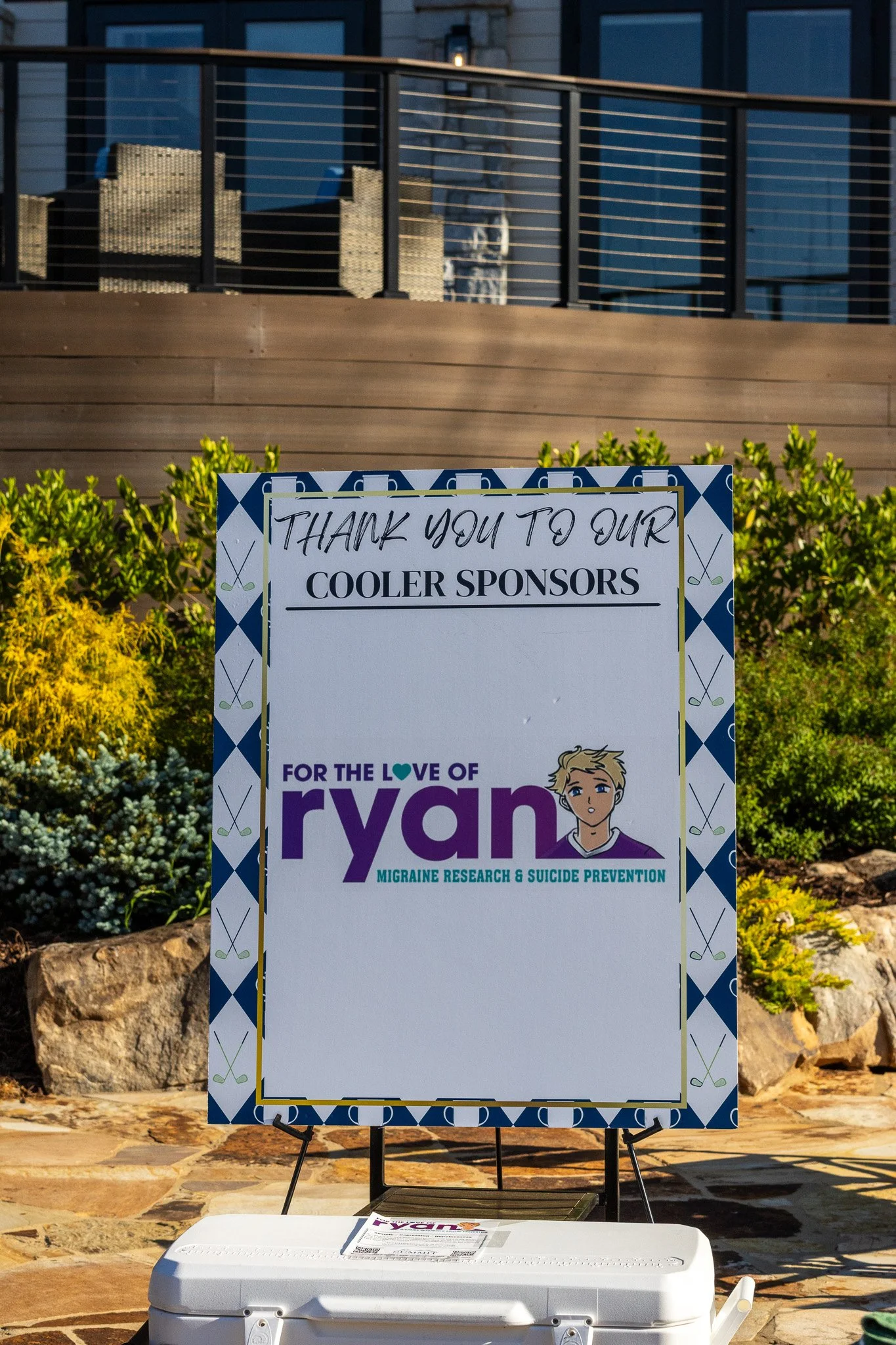 A sign thanking donors for supporting Ryan's migraine research and suicide prevention, with a cartoon image of Ryan, covered with bushes, rocks, and a building with balcony in the background.