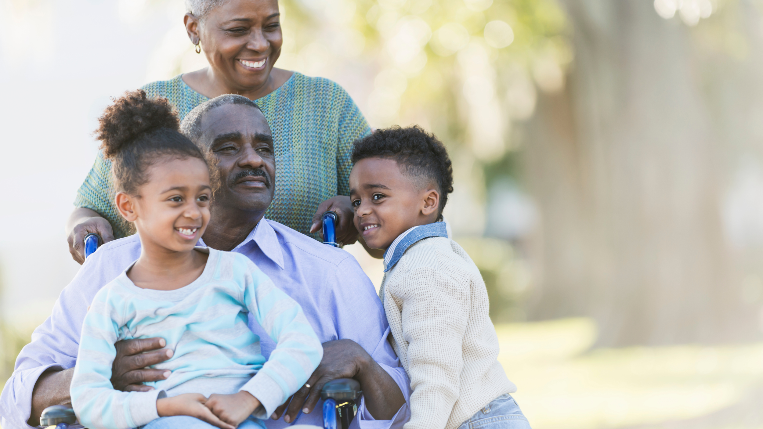 A joyful elderly African American man in a wheelchair is surrounded by a smiling African American woman and two young children as they spend time together outdoors in a park with bright sunlight and green trees in the background.