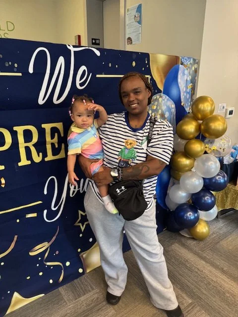 Woman holding a young girl standing in front of a decorated backdrop with gold, silver, and blue balloons.