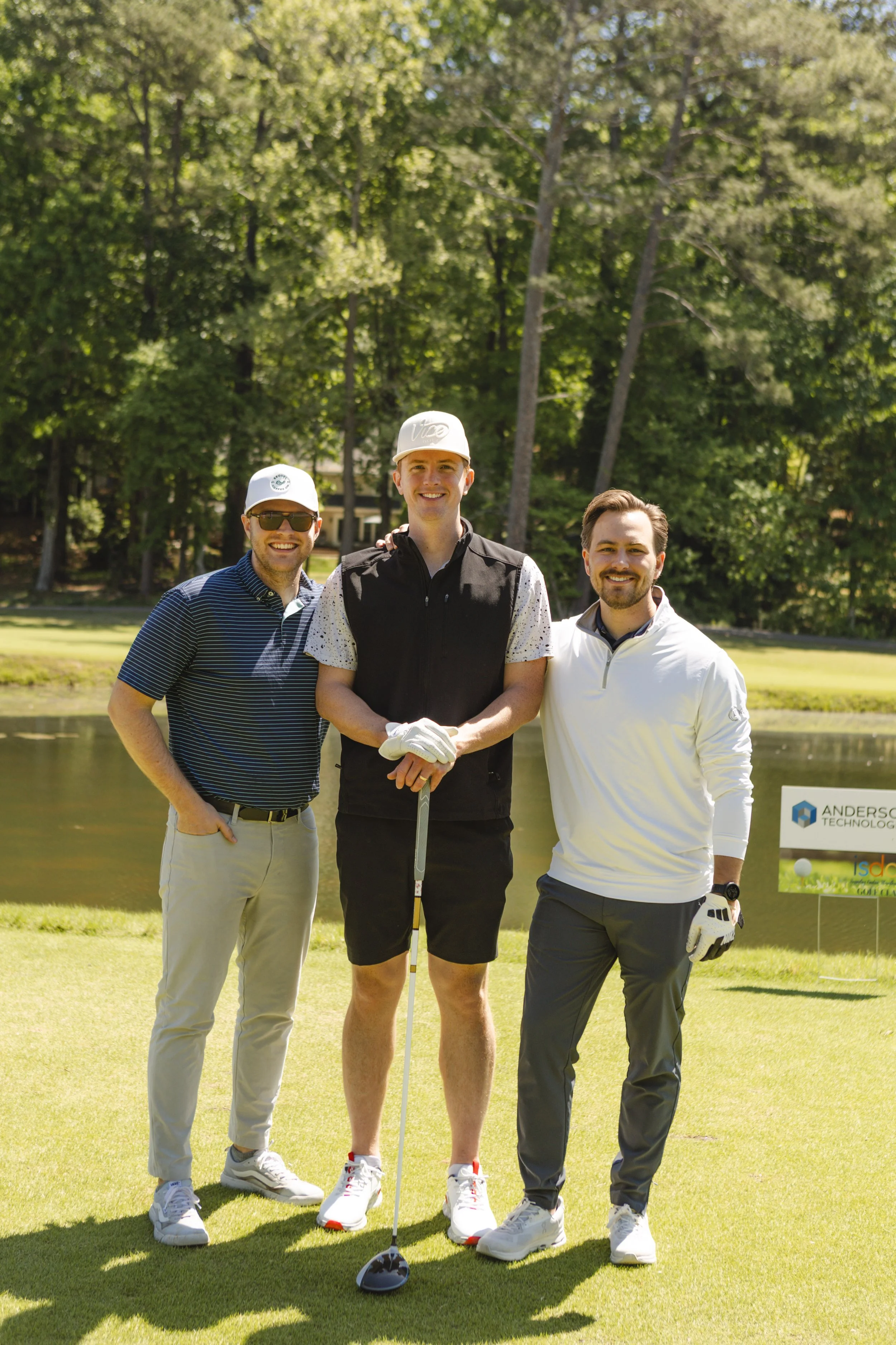 Three men standing on a golf course, smiling at the camera, with trees in the background. One man is holding a golf club, and another has a golf glove. The man in the middle is wearing a white cap and a black vest.