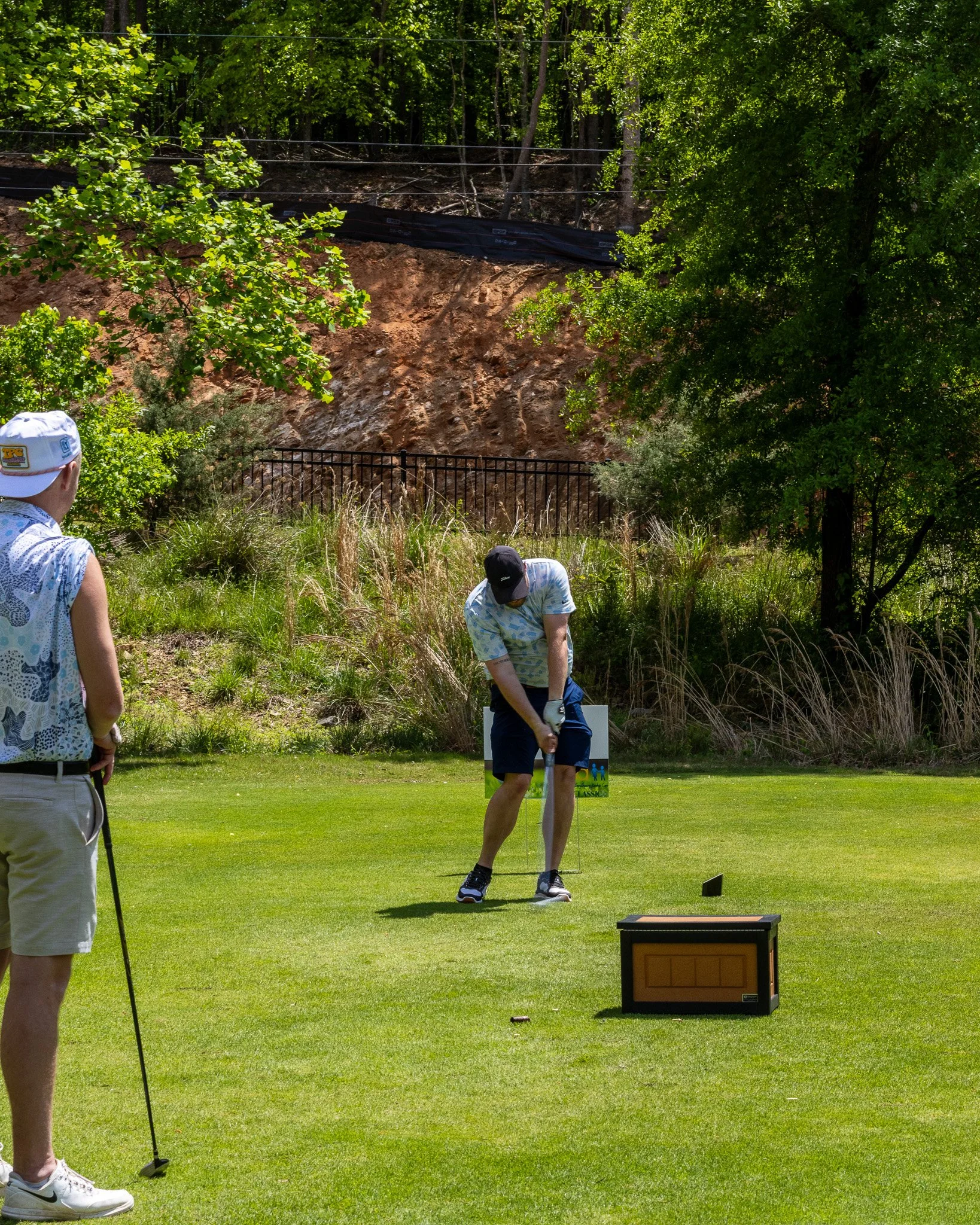 Three men playing mini golf outdoors on a sunny day, one preparing to swing, two others watching, with trees, grass, and a box in the background.