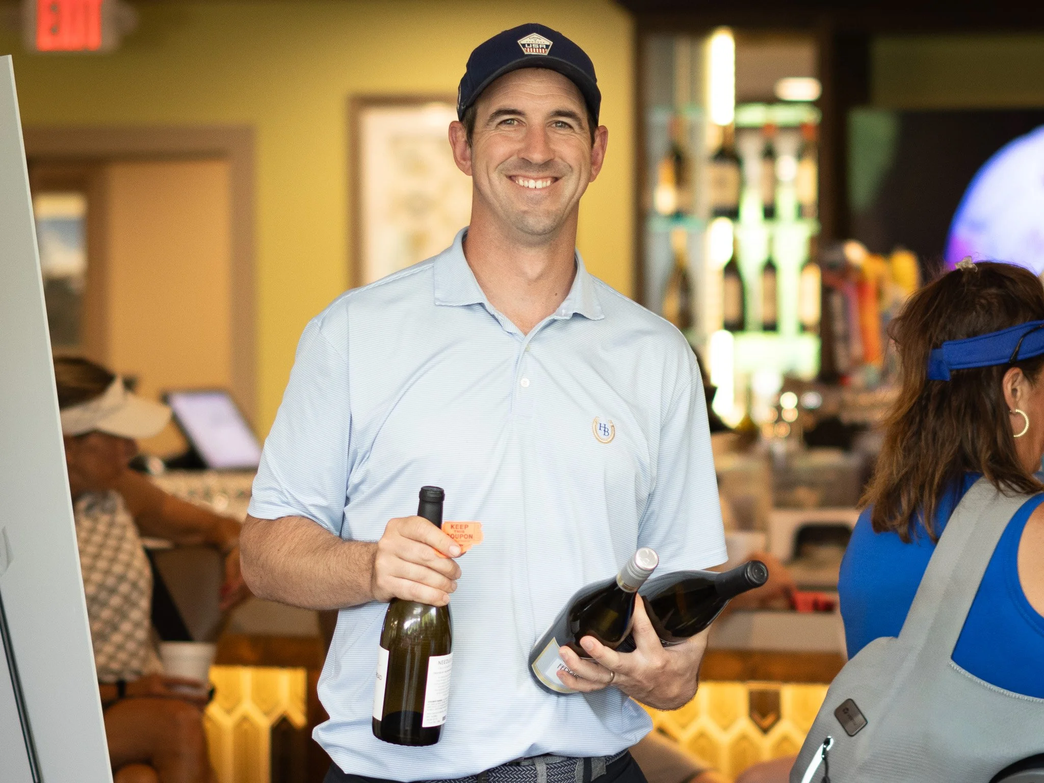 A smiling man in a light blue polo shirt and navy cap with an USA emblem takes a corkscrew and two bottles of wine at a wine tasting event. He holds a corkscrew and a bottle of white wine, standing in a warmly lit venue with shelves and people in the