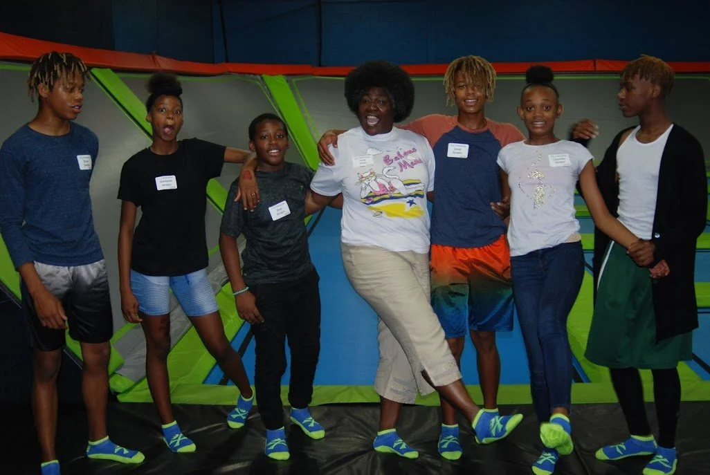 Group of seven children and one woman standing on trampoline with colorful padded wall in indoor trampoline park