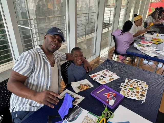 Man and young boy smiling at the camera, sitting at a table with art supplies and cut-out pictures, in a bright indoor space with large windows and other people working on crafts in the background.