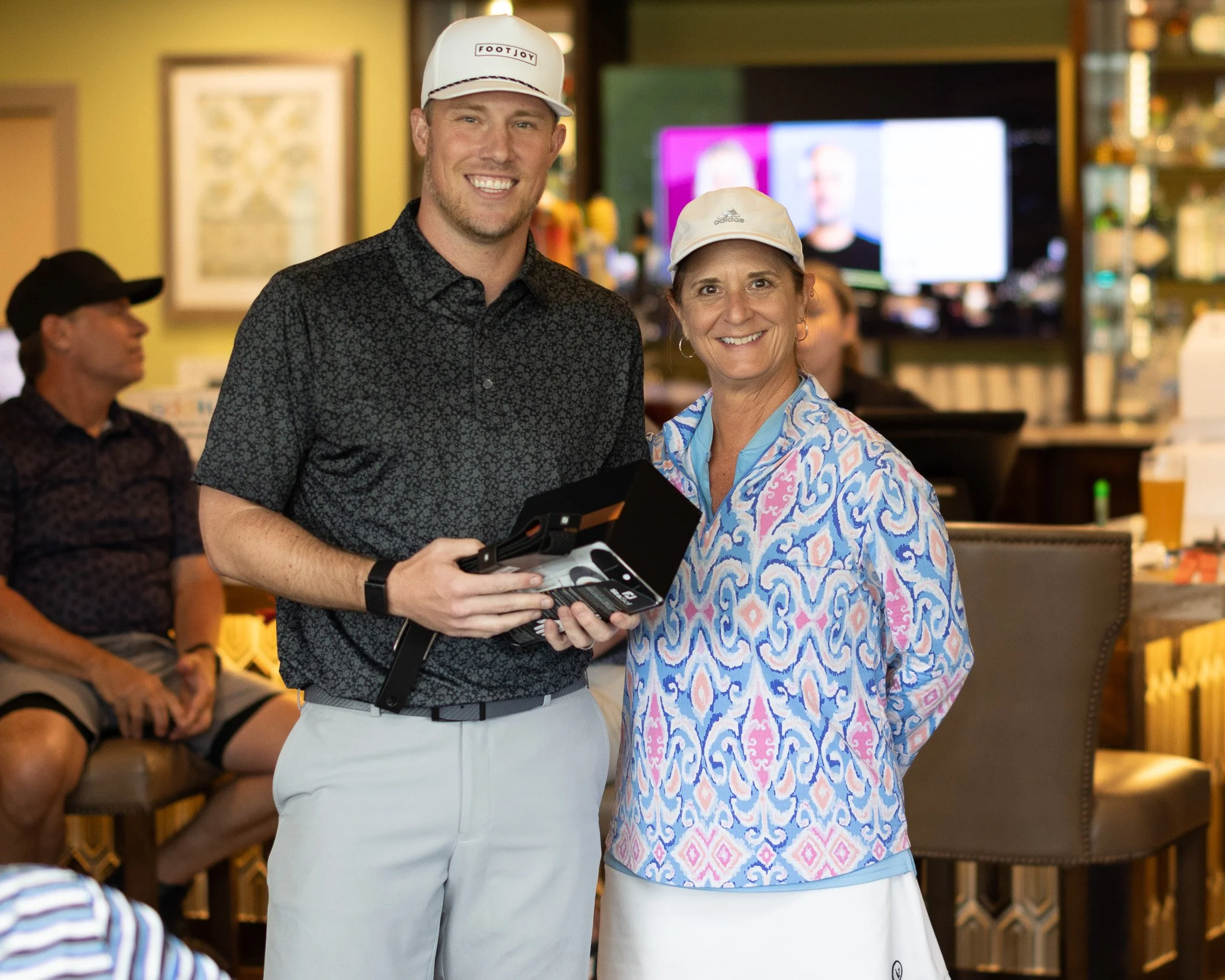 Two smiling people, a man and a woman, stand in a restaurant. The man wears a white cap with a logo, a dark patterned shirt, and light-colored pants. The woman wears a light-colored cap, a colorful patterned top, and has earrings. The background incl