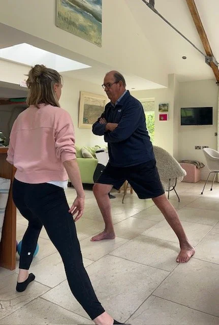 A woman and a man are practicing martial arts in a living room. The woman is standing, and the man, dressed in a blue jacket and shorts, is in a fighting stance with one leg forward and arms crossed.