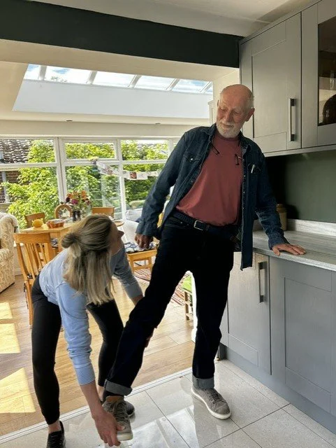 A woman is squatting on the floor tying a man's shoe in a kitchen with natural light and a dining area in the background.