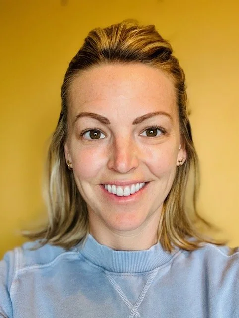 A woman with light brown hair, smiling, wearing a light blue top, with a yellow background.