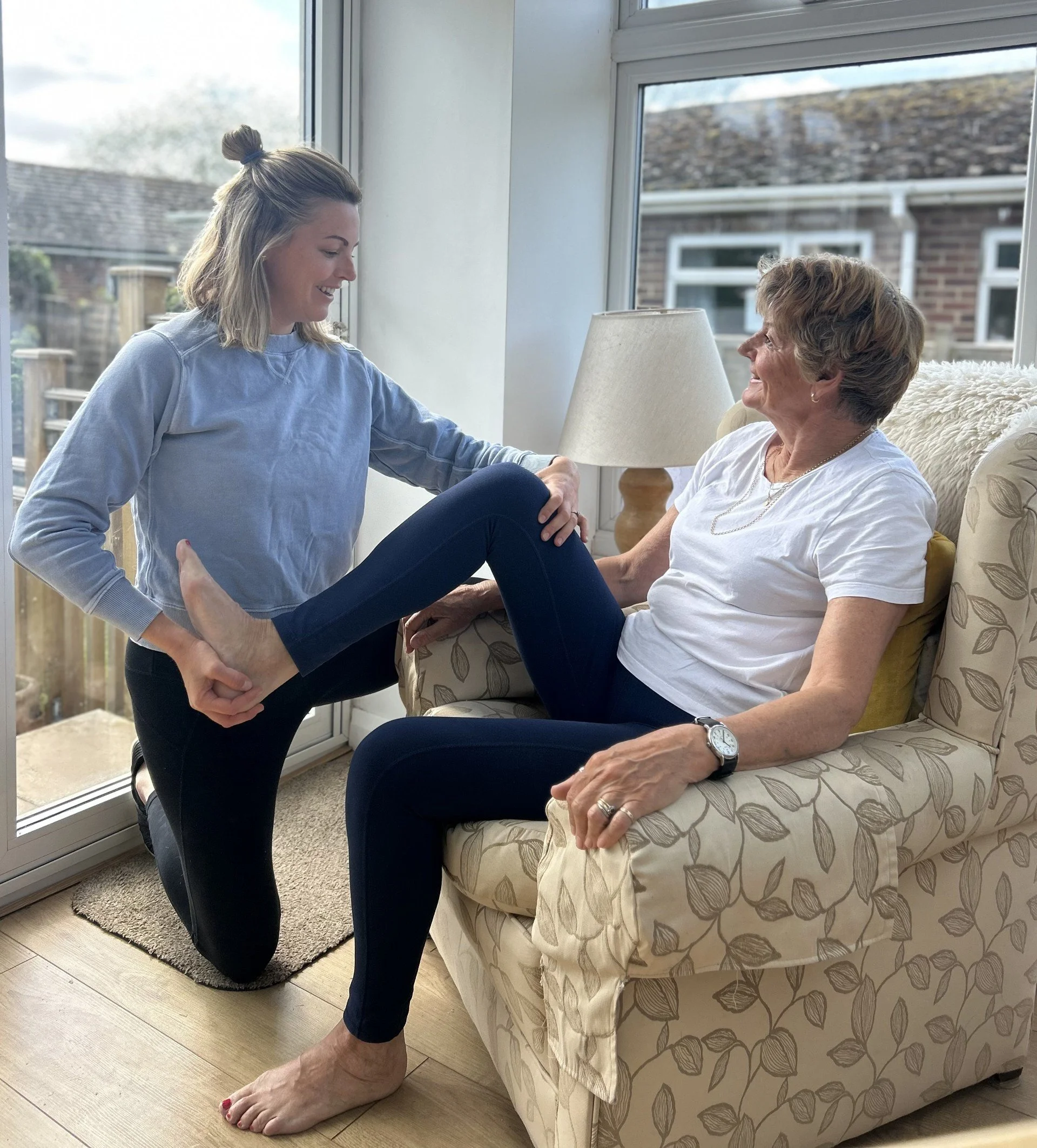 A young woman and an elderly woman smiling together in a bright living room near a window, with the young woman kneeling and holding the elderly woman's leg.