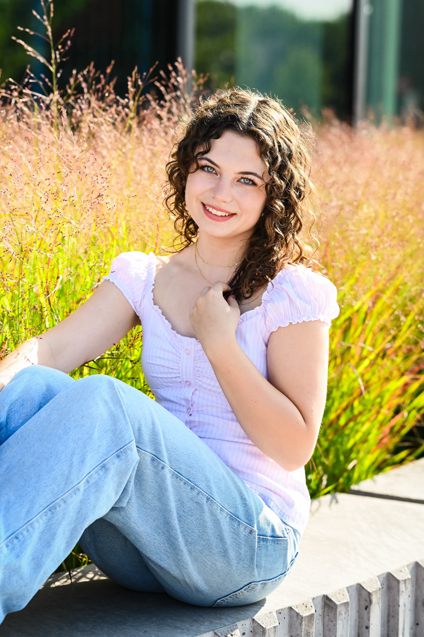 A young woman with curly brown hair and blue eyes smiling, sitting outdoors on a concrete ledge in front of pink and green ornamental grass, wearing a white short-sleeved top and light blue jeans, with sunlight shining on her face.