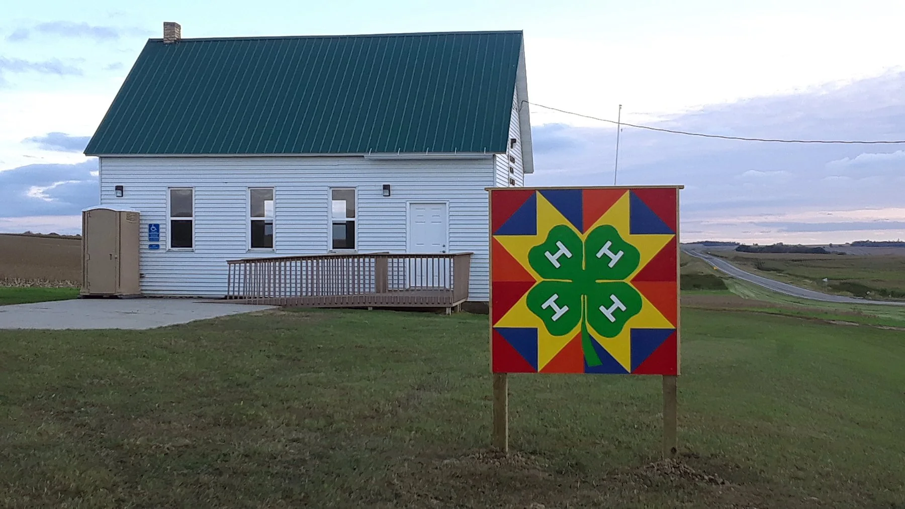 A white building with a green metal roof, surrounded by grass and an open landscape. In front, a colorful art sign featuring a four-leaf clover with four green 'H' symbols, set against a red, yellow, and blue background.