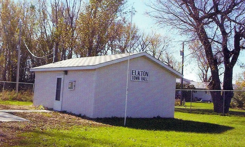 A small white building labeled 'Elkton Town Hall' with a metal roof, located on a grassy area with trees and a chain-link fence in the background.