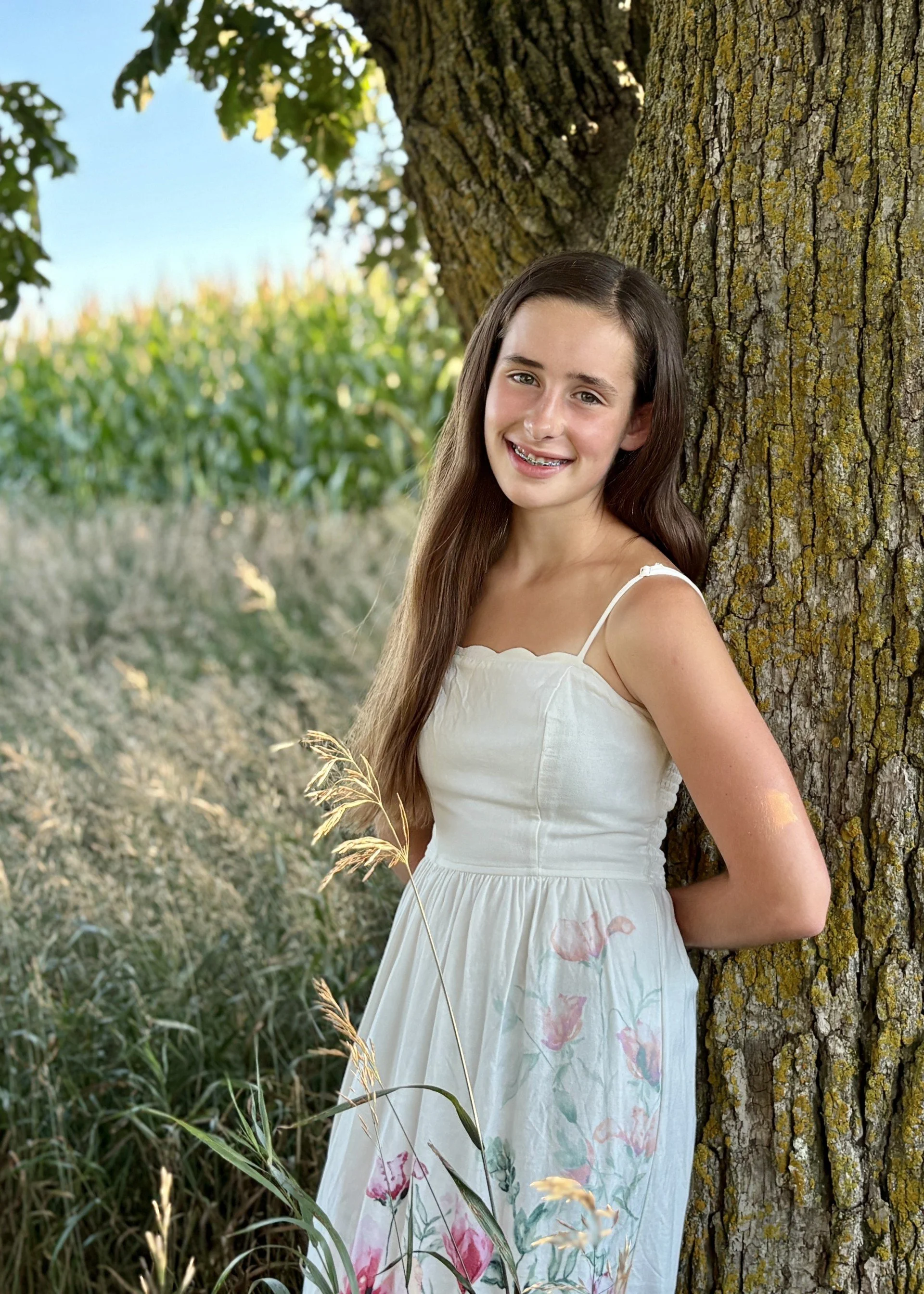A young woman with long brown hair and braces, wearing a white dress with pink and purple floral patterns, leaning against a tree trunk in a grassy outdoor setting, smiling at the camera.