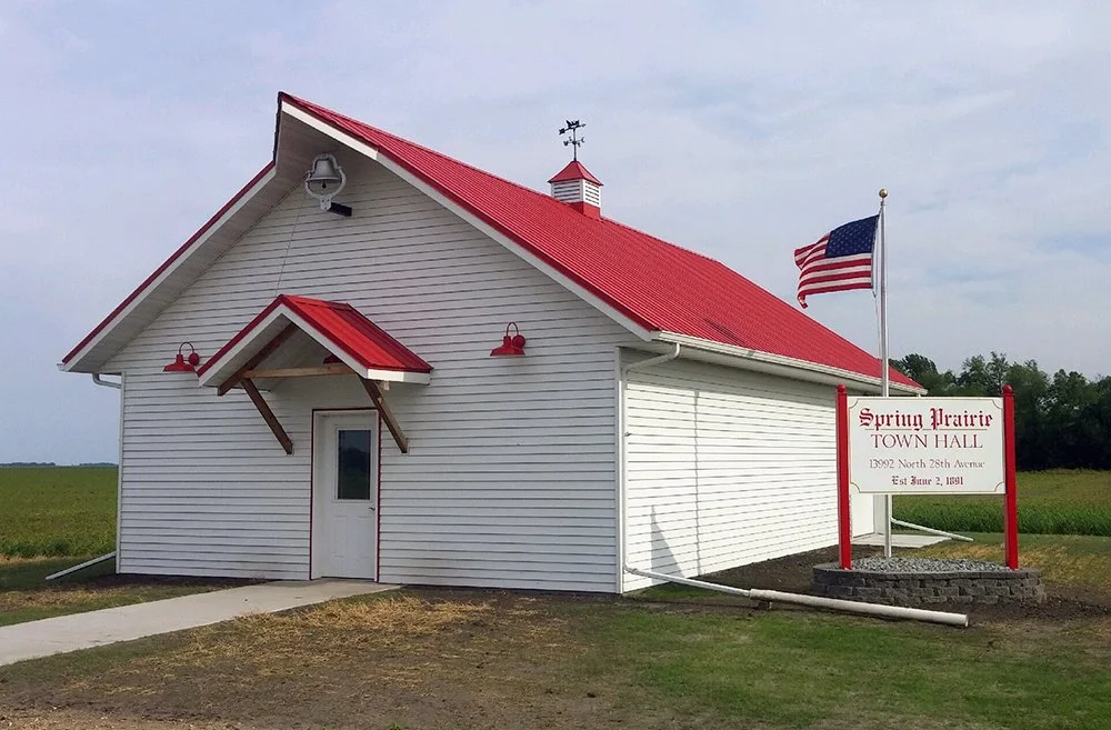 A white town hall building with red roof accents, a small porch, and a sign that reads 'Spring Prairie Town Hall, 1392 North 28th Avenue, Est. June 2, 1891.' An American flag is flying next to the sign.
