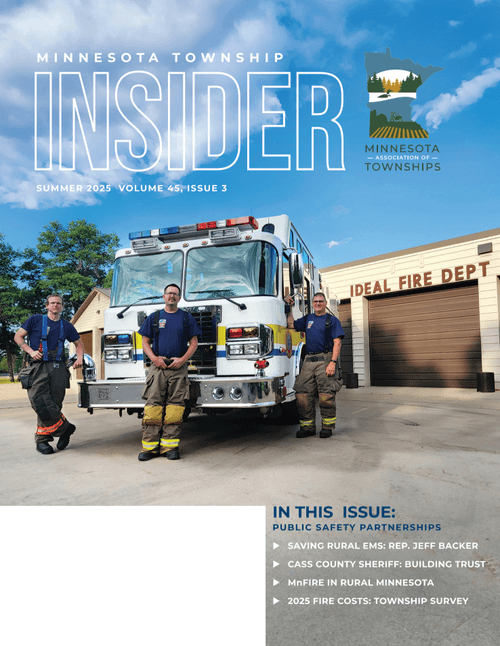 Four firefighters standing in front of a fire truck and fire station with a sign that reads "Ideal Fire Dept." in Minnesota, summer 2025.