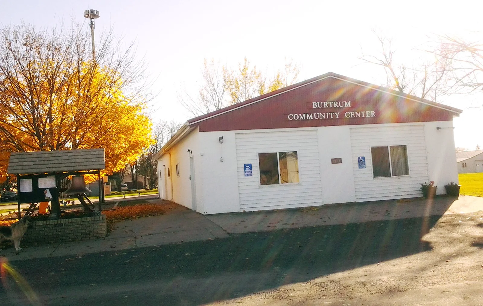 The small white building with a brown gabled roof has a sign reading 'Burtrum Community Center.' There are two blue handicapped parking signs on the front. An autumn tree with yellow leaves is on the left, and a church bell is visible in front of the