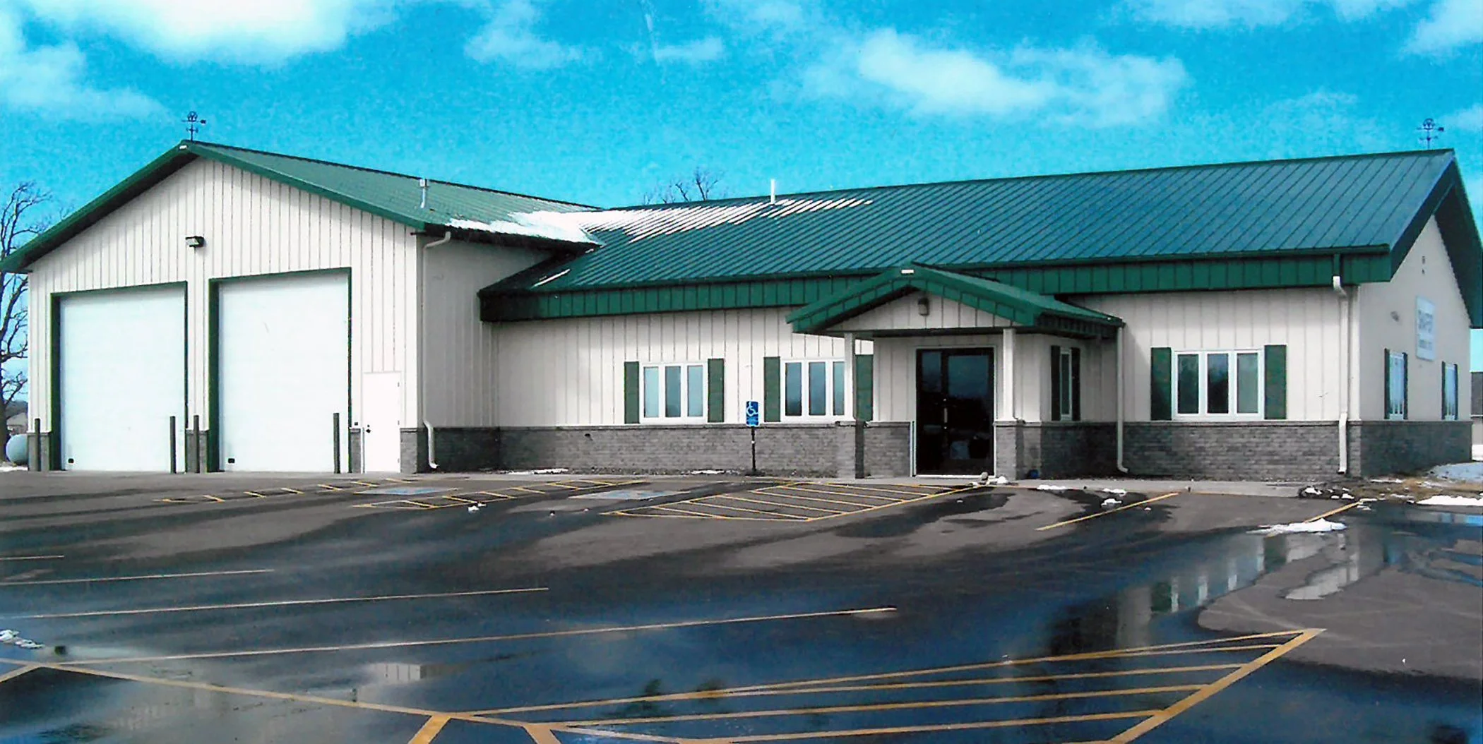 A commercial building with a green metal roof and white siding, featuring two large garage doors, a smaller entrance door, and several windows. The parking lot in front has marked parking spaces and some snow patches, with a partly cloudy sky overhea