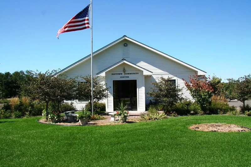 White community center building with a peaked roof, surrounded by grass and bushes, American flag flying on a flagpole in front, under a clear blue sky.