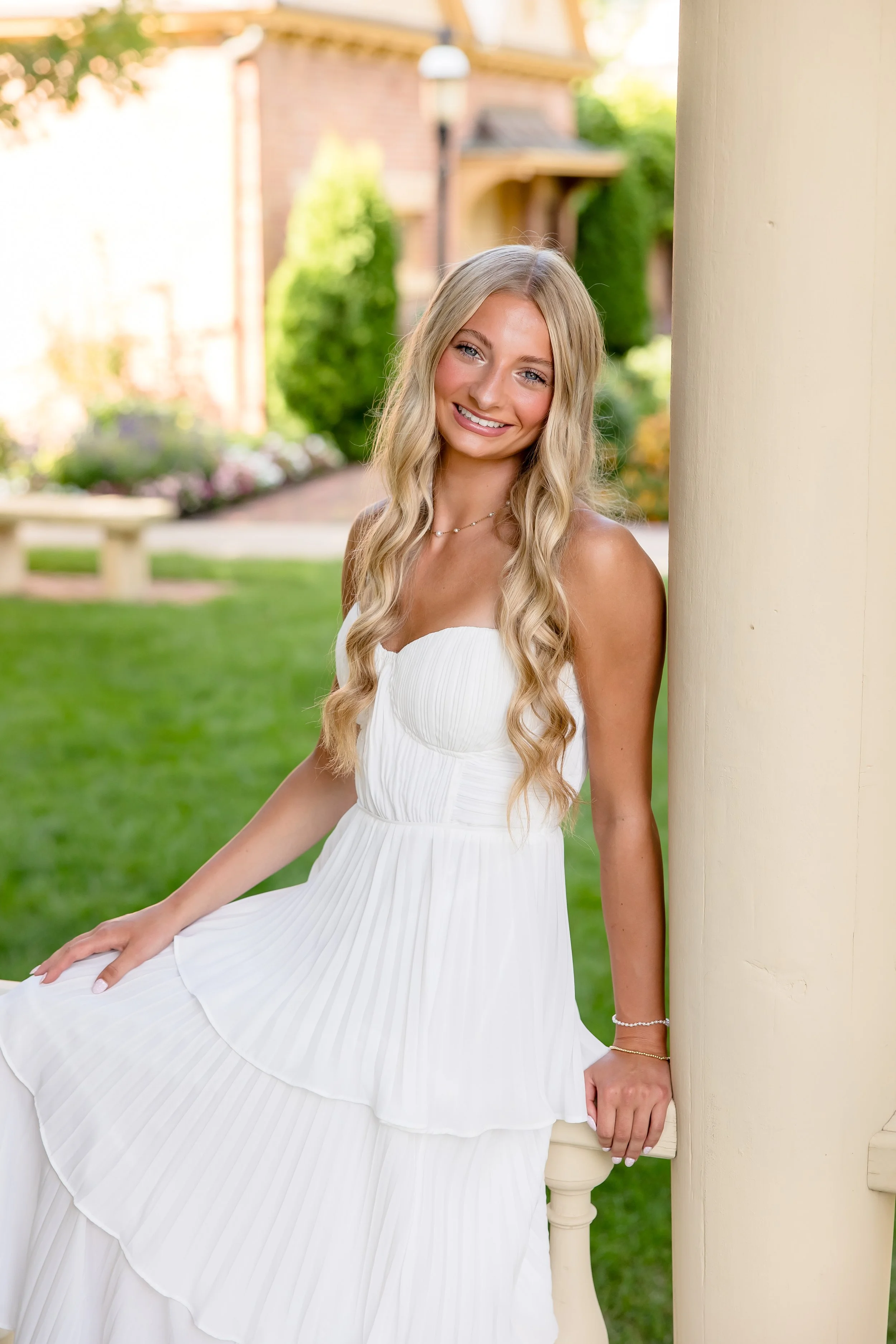 A young woman with long blonde hair, smiling, wearing a white strapless dress, sitting outdoors near a beige column, with a blurred background of trees, bushes, and a brick house.
