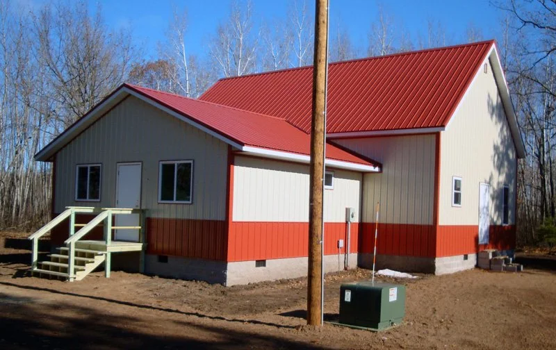 A newly constructed house with beige siding and red accents, a red metal roof, small front porch with stairs, situated on a cleared lot with bare trees in the background.