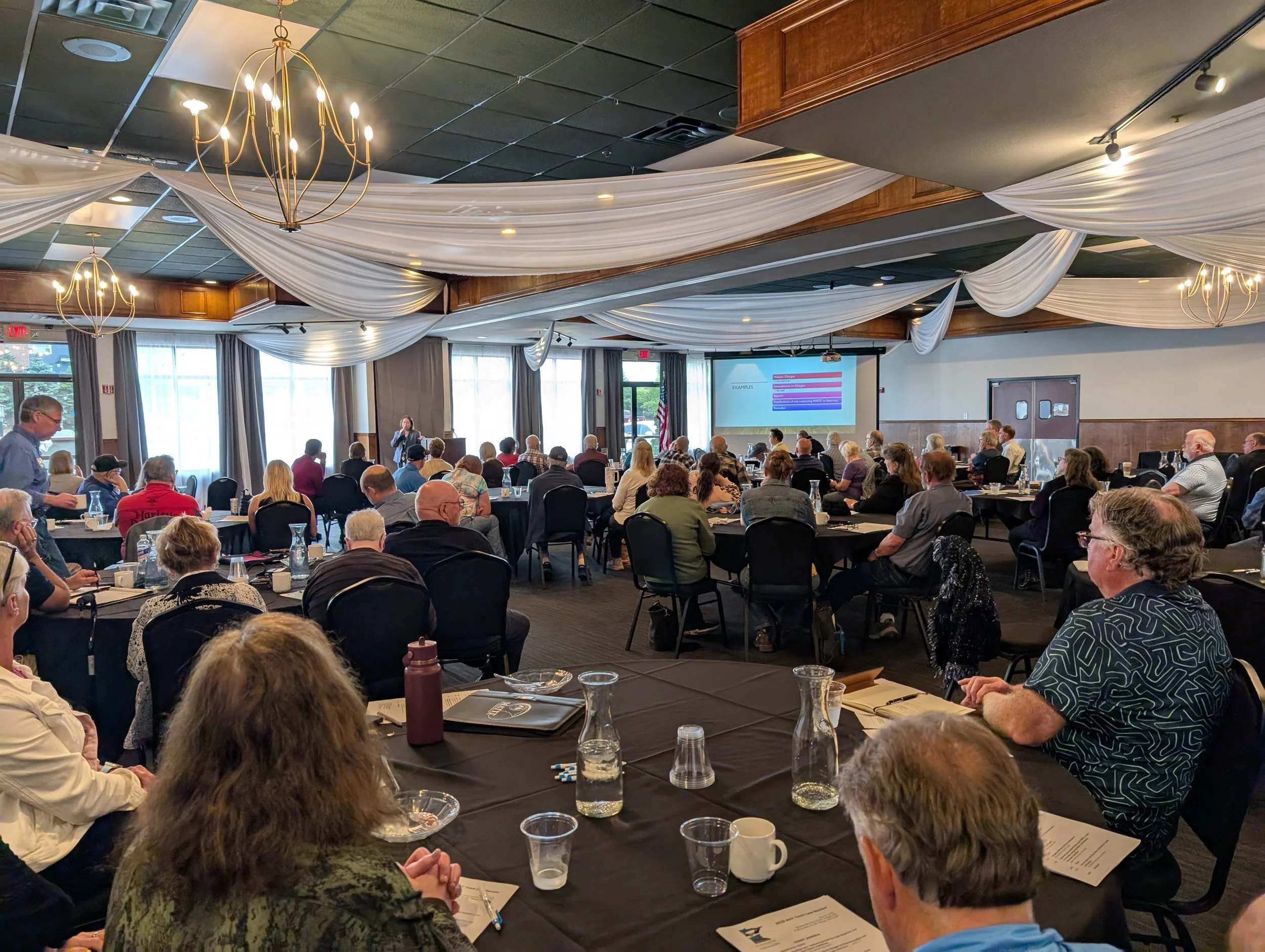 A large conference room or banquet hall filled with people attending a presentation or seminar. There are white decorative drapes or fabric banners hanging from the ceiling, and chandeliers providing lighting. The room has windows with curtains, and a projector screen displays a presentation slide at the front.