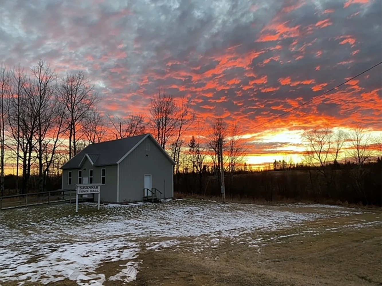 A small gray town hall building with a sign that reads 'Saxenberg Town Hall' is situated in a rural area with a dirt and snow-covered ground. Behind it, leafless trees are silhouetted against a dramatic sunset sky with vibrant orange, pink, and purpl