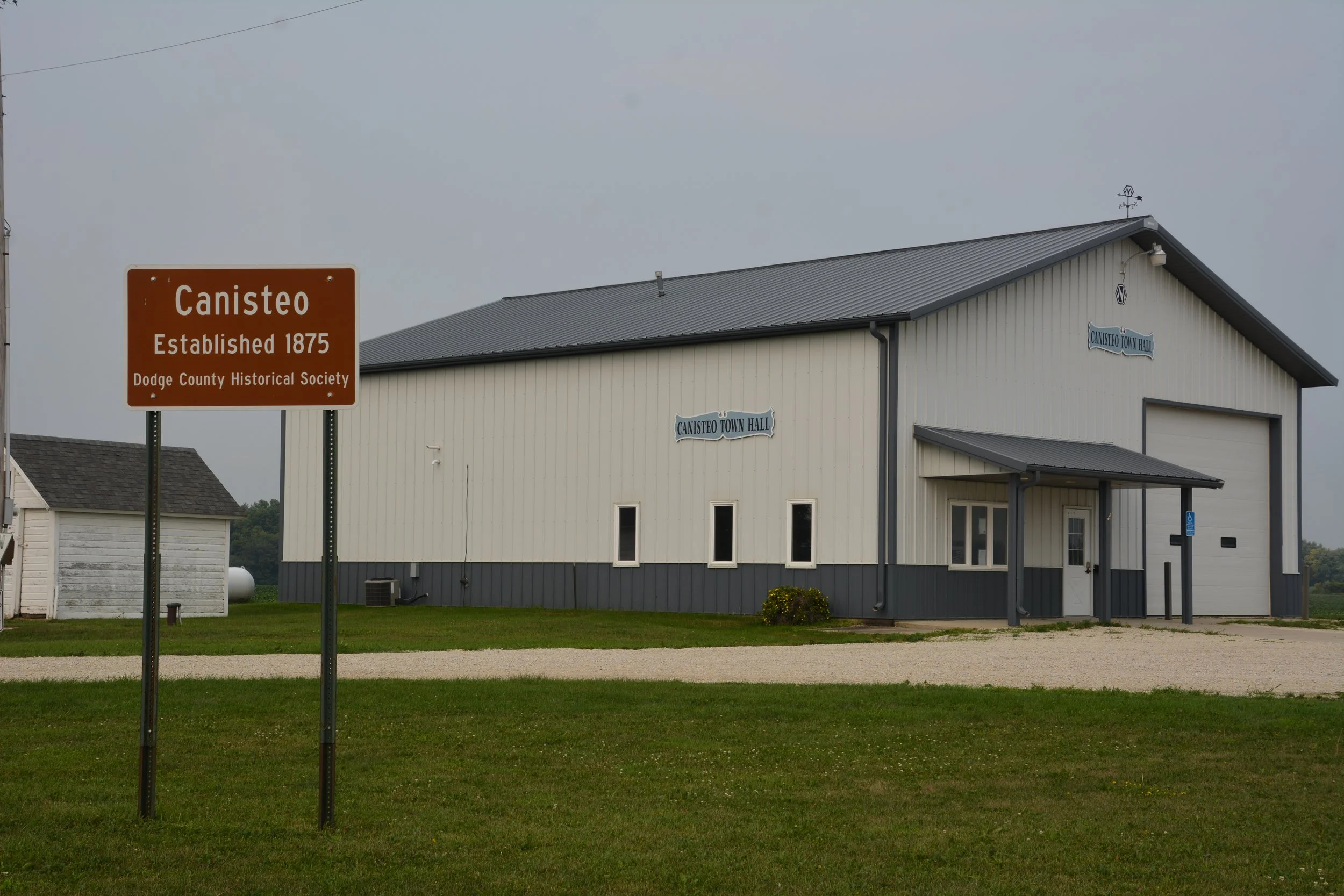 A beige and gray building with a sign reading 'Canisteo Town Hall' and another sign indicating 'Established 1875' by the Dodge County Historical Society. The building is situated on a grassy area with a gravel path in front, under a cloudy sky.