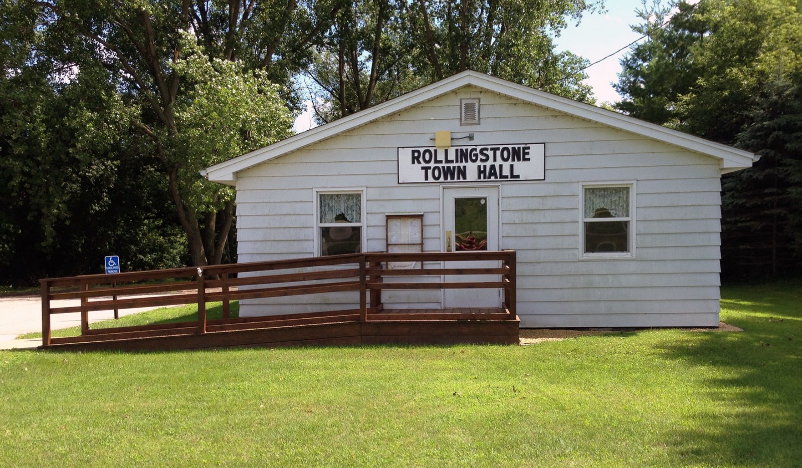 A small white building with a sign that reads "Rollingstone Town Hall" in black letters. There is a wooden ramp leading to the front door. The building is surrounded by green grass and trees.