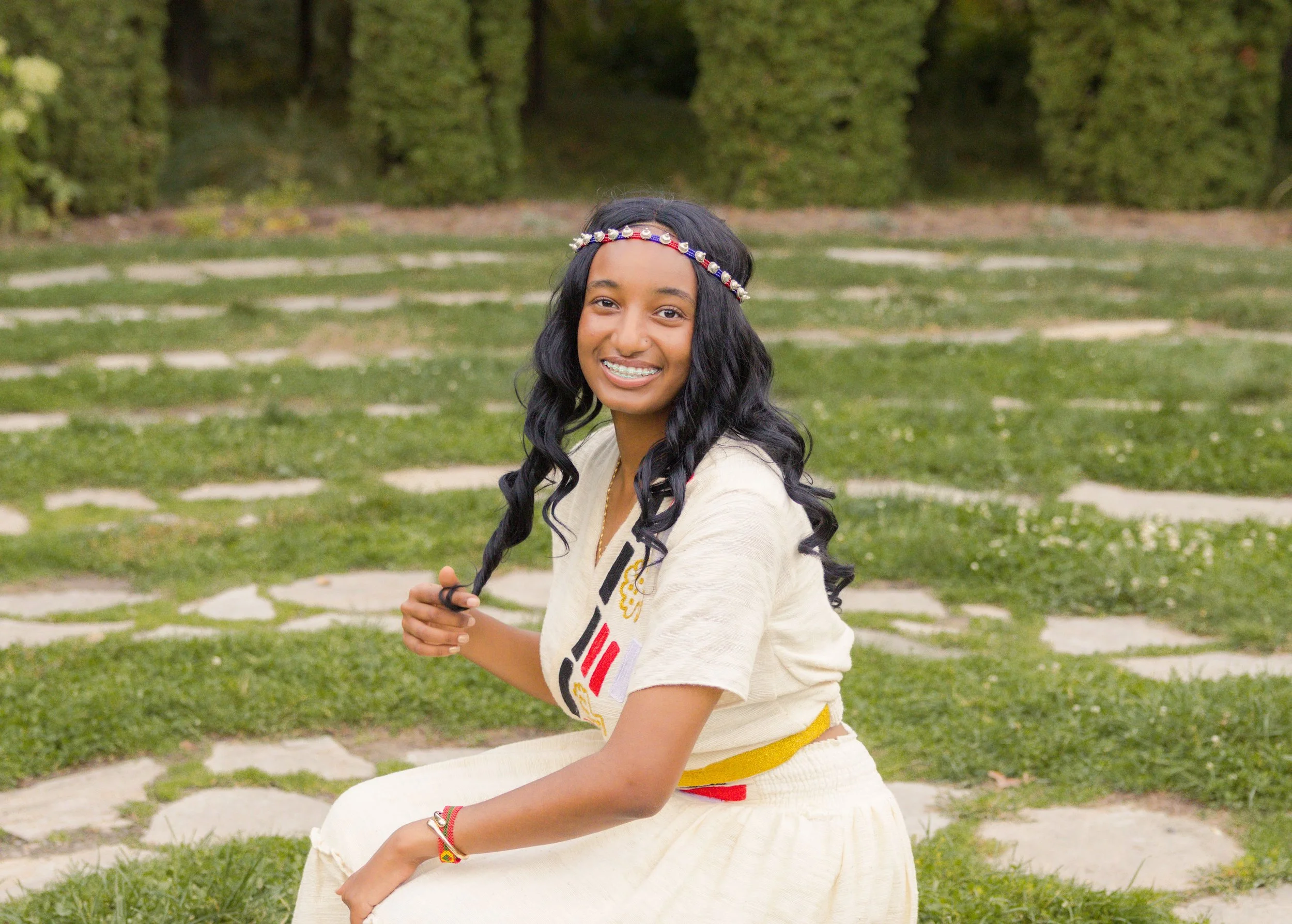 A smiling woman with long black hair, wearing a colorful headband and a cream-colored top, sitting on a stone bench in a park with green grass and trees in the background.