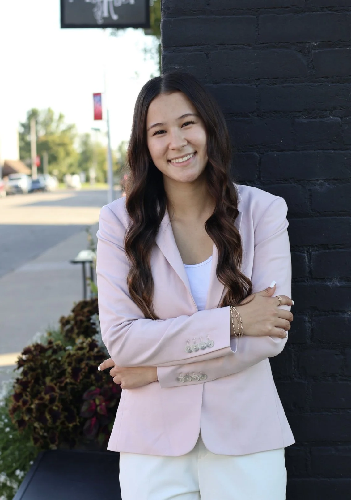 Young woman with long wavy dark hair smiling, wearing a light pink blazer over a white top, standing outdoors against a black brick wall with a blurred street background.