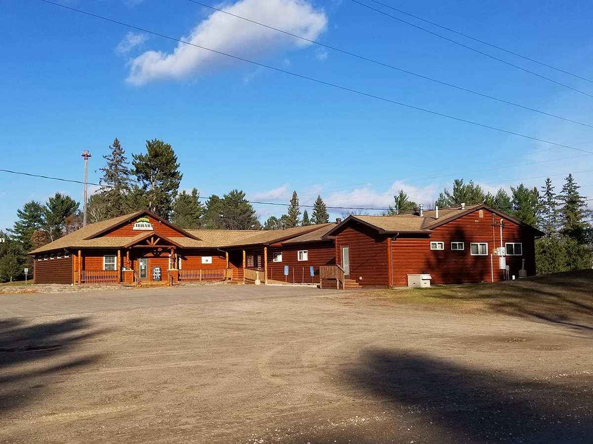 A large wooden building with a sign that reads 'Library,' surrounded by trees and a mostly empty dirt parking lot under a clear blue sky with a few clouds.