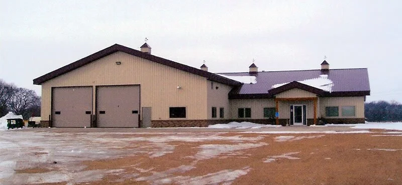 A large beige building with a metal roof, two garage doors, and several small windows, surrounded by snow and a wide open area.