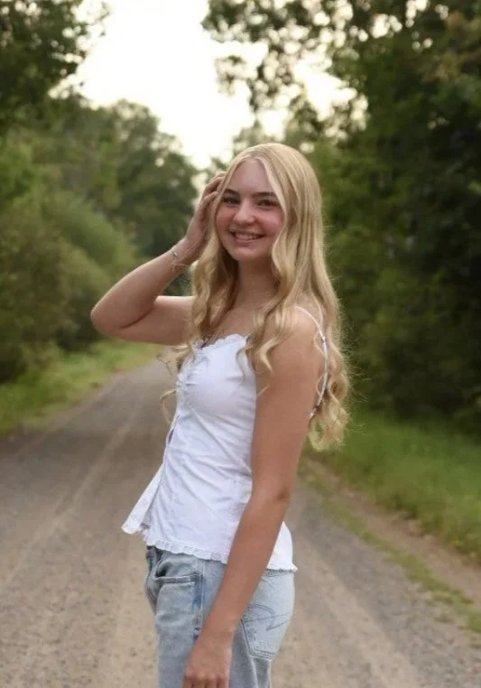 A young woman with long blonde hair smiling and standing on a dirt path in a wooded area.