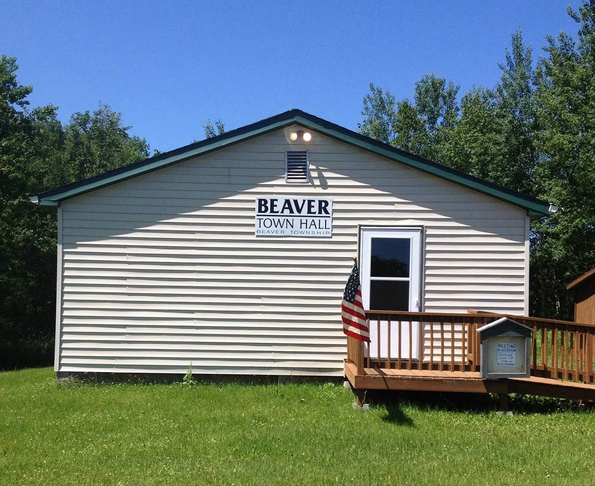 A small beige building labeled Beaver Town Hall with a sign reading 'Beaver Township.' There is a wooden porch with an American flag and a bulletin board, and trees are in the background.