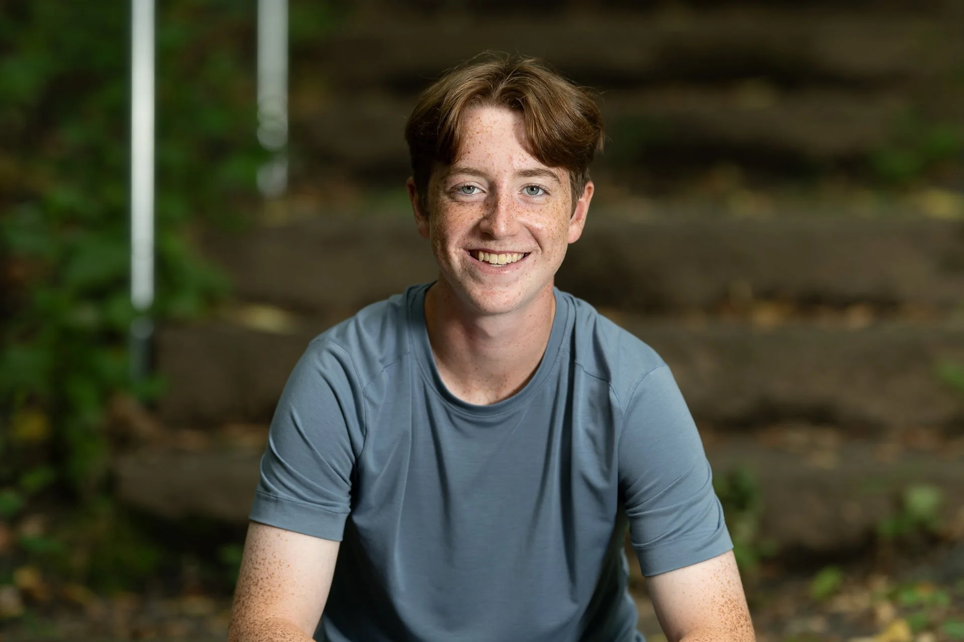A young man with red hair and freckles smiling while sitting outdoors on a trail surrounded by greenery.