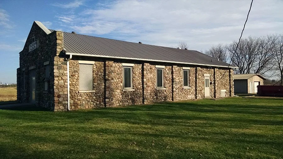A stone building with a metal roof, four windows, and a white front door on a grassy field with a small outbuilding and trees in the background.
